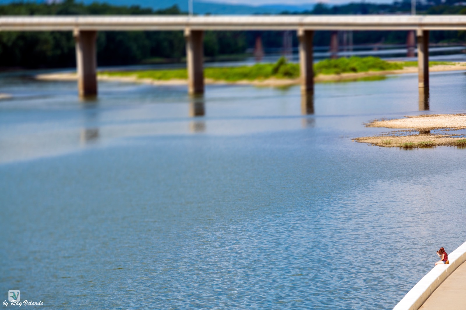 A solitary person sitting beside a broad river beneath a distant bridge