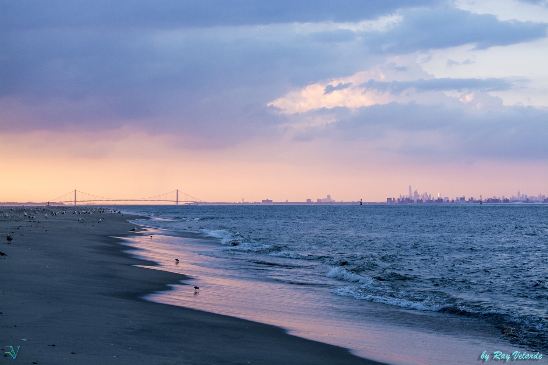 Shoreline, bridge, and New York City skyline beneath dramatic pastel clouds